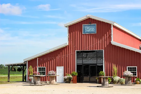Exterior view of a red metal barn wedding venue with a sign reading 'Myers Wedding Barn', decorated with string lights and floral arrangements on wooden pedestals.