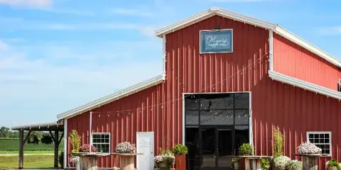 Exterior view of a red metal barn wedding venue with a sign reading 'Myers Wedding Barn', decorated with string lights and floral arrangements on wooden pedestals.
