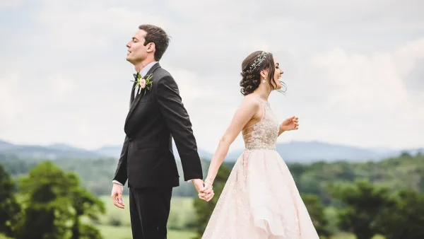 A bride with a styled updo and floral headpiece walking hand-in-hand with a groom in a scenic outdoor setting.