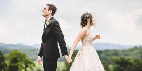 A bride with a styled updo and floral headpiece walking hand-in-hand with a groom in a scenic outdoor setting.