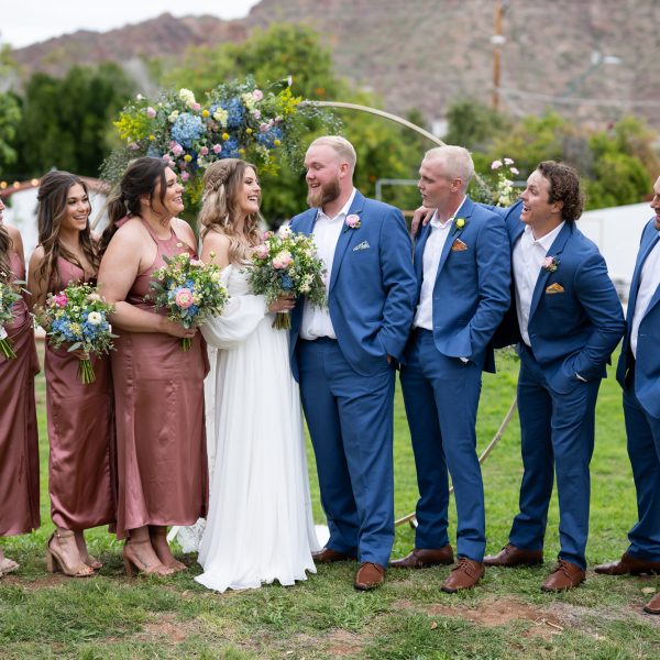 A bride and groom in formal attire standing with their wedding party outdoors on a grassy lawn with mountains in the background.
