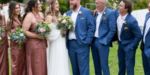 A bride and groom in formal attire standing with their wedding party outdoors on a grassy lawn with mountains in the background.