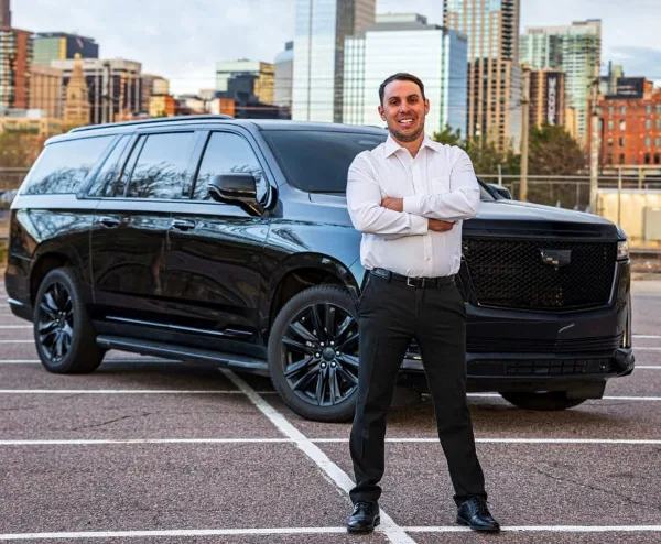 A professional chauffeur in a white shirt and black trousers stands with arms crossed in front of a black Cadillac Escalade SUV in a parking lot with a city skyline in the background.