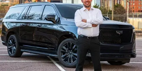 A professional chauffeur in a white shirt and black trousers stands with arms crossed in front of a black Cadillac Escalade SUV in a parking lot with a city skyline in the background.