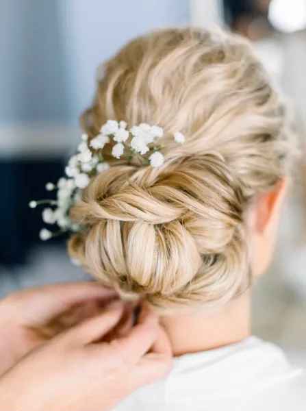 A close-up of a hairstylist's hands finishing a braided updo with white flowers on a client.