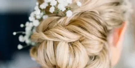 A close-up of a hairstylist's hands finishing a braided updo with white flowers on a client.