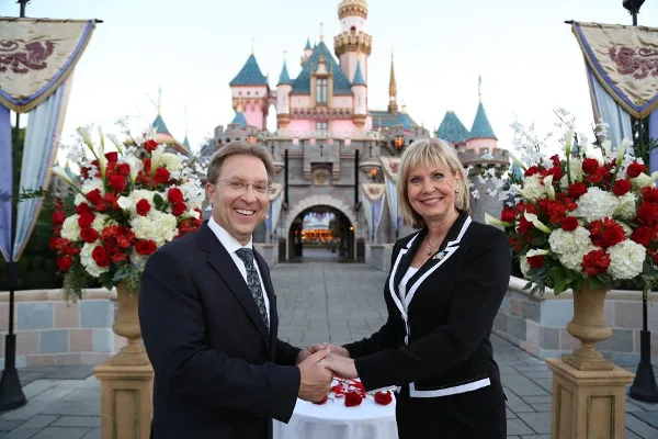 A man and a woman in formal black attire exchange rings at a small table with red rose petals in front of a castle.