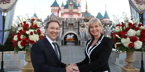 A man and a woman in formal black attire exchange rings at a small table with red rose petals in front of a castle.