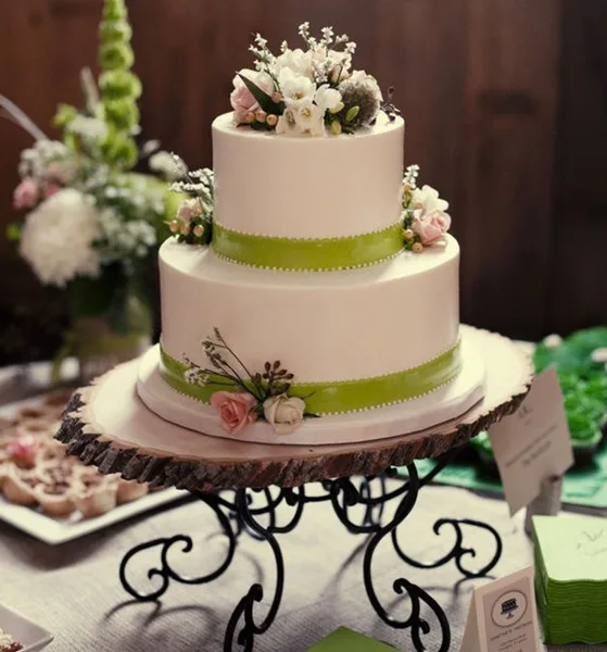 A two-tier white wedding cake with green ribbon accents and floral decorations on a wooden stand.