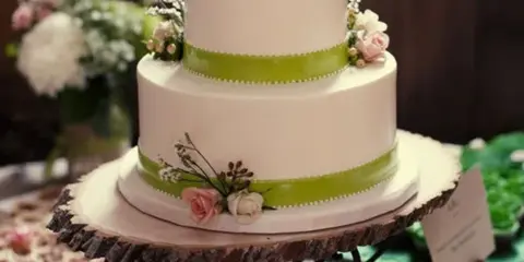 A two-tier white wedding cake with green ribbon accents and floral decorations on a wooden stand.