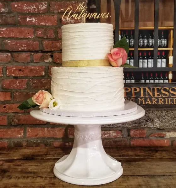 A two-tier white wedding cake with textured frosting, decorated with pink roses and a gold ribbon, displayed on a white ceramic stand.