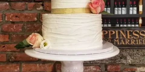 A two-tier white wedding cake with textured frosting, decorated with pink roses and a gold ribbon, displayed on a white ceramic stand.
