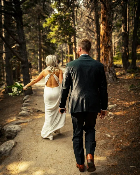 A bride and groom walking away from the camera on a forest path, with the bride holding a bouquet.