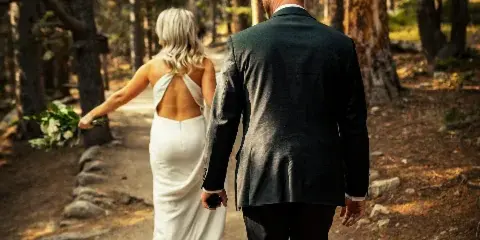 A bride and groom walking away from the camera on a forest path, with the bride holding a bouquet.