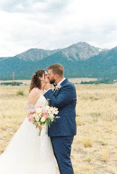 A bride and groom kissing in a grassy field with a large mountain range in the background.