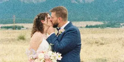 A bride and groom kissing in a grassy field with a large mountain range in the background.