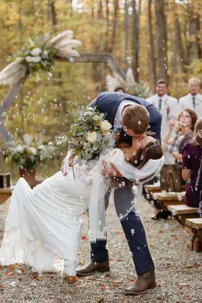 A groom dips his bride for a kiss during an outdoor wedding ceremony in a wooded area.