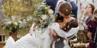 A groom dips his bride for a kiss during an outdoor wedding ceremony in a wooded area.