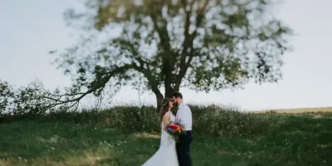 A bride and groom standing together in a grassy field with a large tree in the background.