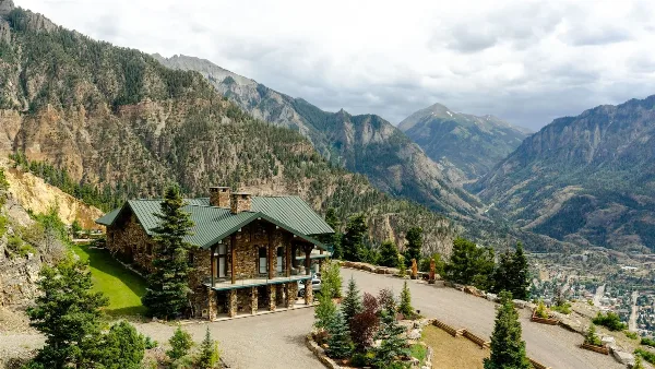Aerial view of a stone lodge with a green roof and stone chimney, situated on a mountain ridge with a valley and town visible in the distance.