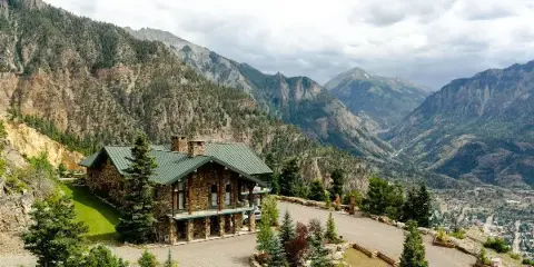 Aerial view of a stone lodge with a green roof and stone chimney, situated on a mountain ridge with a valley and town visible in the distance.