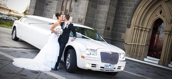 A bride and groom kissing next to a white stretch limousine parked outside a stone building.