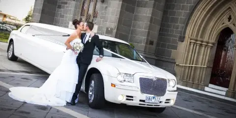 A bride and groom kissing next to a white stretch limousine parked outside a stone building.