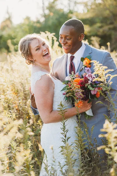 A bride and groom laughing together in a field of tall grass, with the bride holding a colorful bouquet.