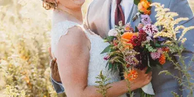 A bride and groom laughing together in a field of tall grass, with the bride holding a colorful bouquet.