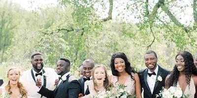 A wedding party including the bride and groom posing outdoors on a grassy lawn with large trees in the background.