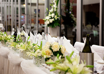 A long banquet table set with white linens, glassware, and floral centerpieces in a bright event venue.