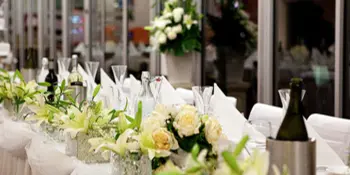 A long banquet table set with white linens, glassware, and floral centerpieces in a bright event venue.