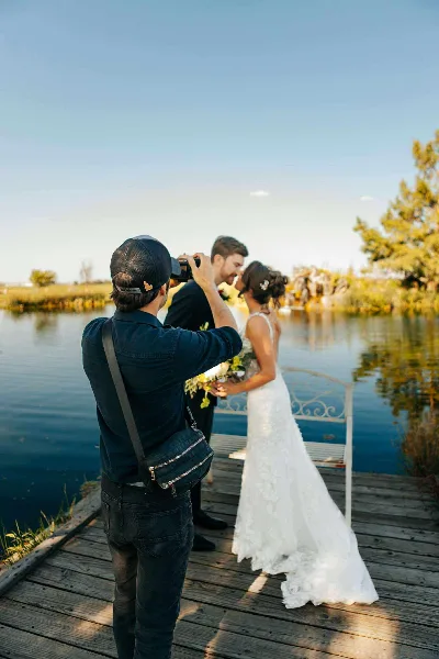 A videographer filming a bride and groom on a wooden dock by a lake.