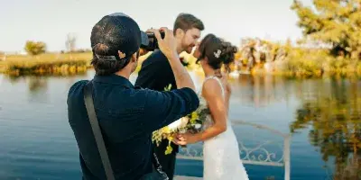 A videographer filming a bride and groom on a wooden dock by a lake.