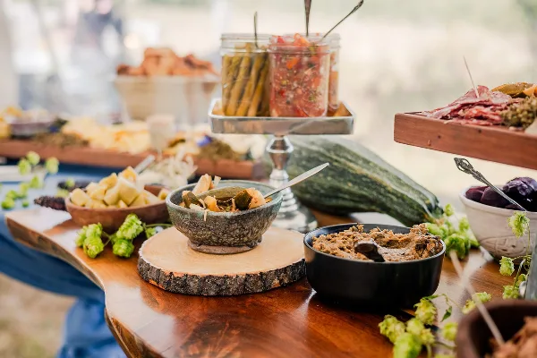 A rustic catering table featuring bowls of seasonal vegetables, dips, and charcuterie arranged on wooden boards and platters.