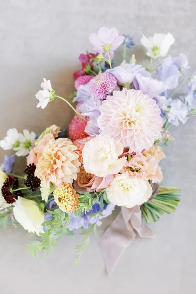 A close-up photograph of a lush floral arrangement featuring pink and peach dahlias, white ranunculus, and greenery, tied with a beige ribbon.