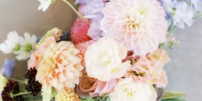 A close-up photograph of a lush floral arrangement featuring pink and peach dahlias, white ranunculus, and greenery, tied with a beige ribbon.