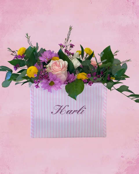 A floral arrangement featuring pink roses, purple daisies, and yellow chrysanthemums in a pink and white striped gift box with the name 'Karli' on the front.