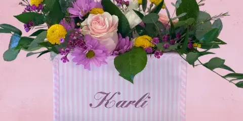 A floral arrangement featuring pink roses, purple daisies, and yellow chrysanthemums in a pink and white striped gift box with the name 'Karli' on the front.