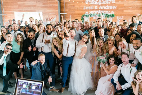 A large group of wedding guests and the couple posing together at a reception with a DJ mixing console visible in the foreground.