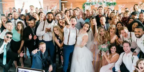 A large group of wedding guests and the couple posing together at a reception with a DJ mixing console visible in the foreground.