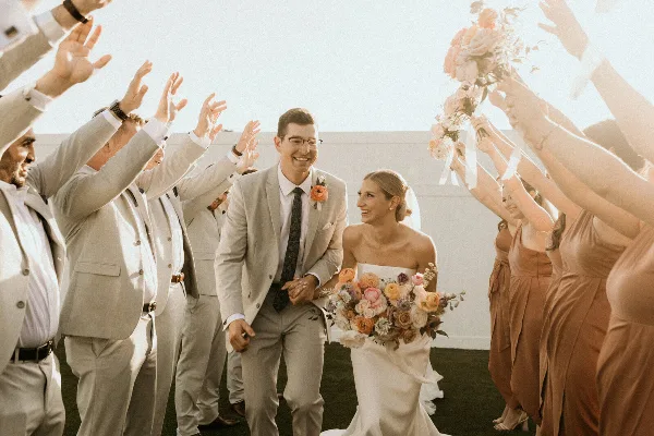 A bride and groom smiling and walking hand-in-hand through a line of cheering wedding guests holding bouquets.