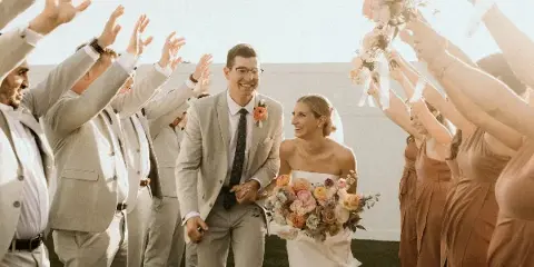 A bride and groom smiling and walking hand-in-hand through a line of cheering wedding guests holding bouquets.