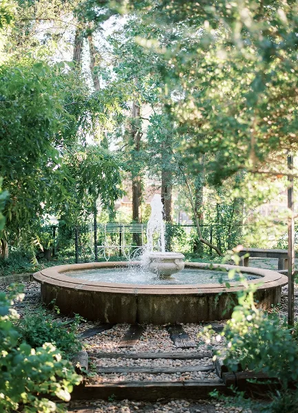 A stone water fountain in a lush, green courtyard garden with trees and a white bench in the background.