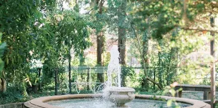 A stone water fountain in a lush, green courtyard garden with trees and a white bench in the background.