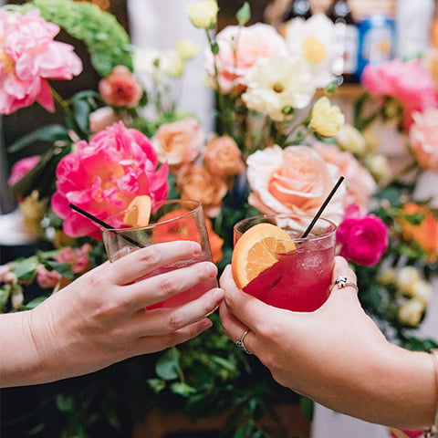 Two hands clinking glasses of pink cocktails garnished with orange slices in front of a floral wedding backdrop.