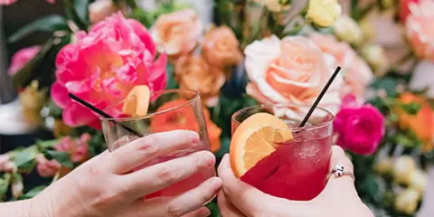 Two hands clinking glasses of pink cocktails garnished with orange slices in front of a floral wedding backdrop.