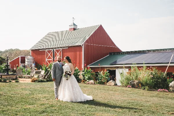 A bride and groom standing on a grassy lawn in front of a large red barn with a water fountain and flower beds.