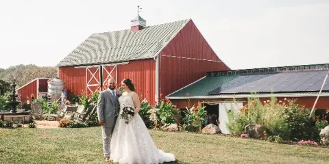 A bride and groom standing on a grassy lawn in front of a large red barn with a water fountain and flower beds.