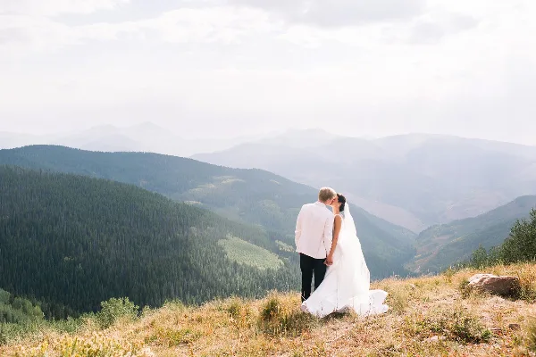 A bride and groom standing on a grassy mountain ridge overlooking a vast valley and forested peaks.